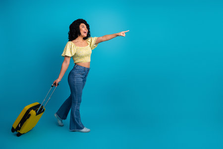 Joyful young woman pointing excitedly while holding a yellow suitcase against a vibrant blue backdropの写真素材