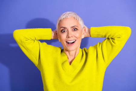 Happy elderly woman in bright yellow sweater poses with hands behind head against blue background...