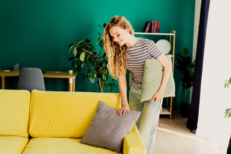 A cheerful young woman tidies a bright living room with a yellow sofa and green wall showcasing a cozy home interior.の写真素材