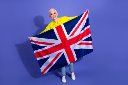 Elderly woman with short blonde hair smiles proudly while holding a Union Jack flag against a vibrant purple background in a bright casual styleの写真素材