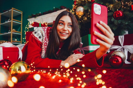 Playful holiday moment as a young woman takes a selfie by a Christmas tree surrounded by gifts and lights in a cozy festive living roomの写真素材
