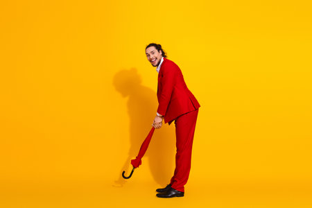 Playful handsome man in a bright red suit smiles while holding a red umbrella against a vivid yellow background for bold fashion and party ready advertisingの写真素材