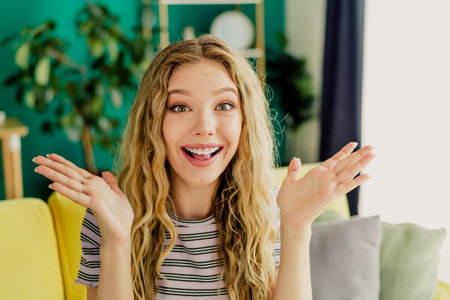 Joyful young woman at home smiles and waves in a cozy living room with bright decor and a colorful couch warm friendly momentの写真素材