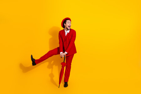 Funny red suit man kicks on bright yellow background wearing hat and holding umbrella for bold stylish fashion photoの写真素材
