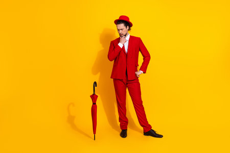 Playful dapper man in red suit and hat strikes a thoughtful pose with a red umbrella against a vivid yellow background creating a bold stylish momentの写真素材