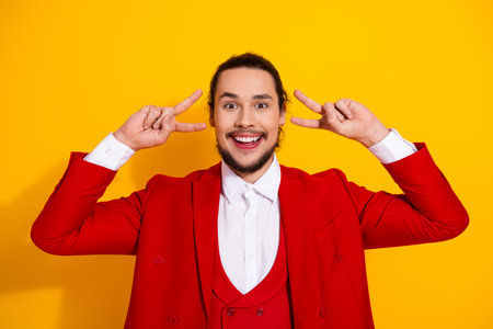 Playful stylish man in red suit says hello with peace signs on a bright yellow backdrop for bold fashion and fun advertisingの写真素材