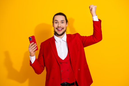 Funny confident man in a bright red suit celebrates a stylish moment on a bold yellow backdrop with a phone in hand and a joyful poseの写真素材