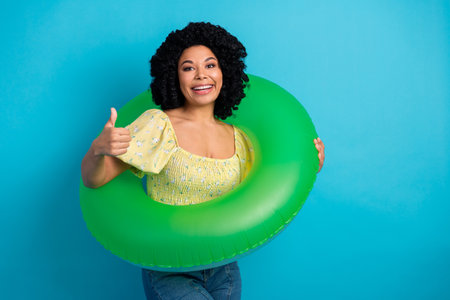 Attractive young woman with curly hair in casual yellow top holding a green float and showing thumbs up against blue backgroundの写真素材