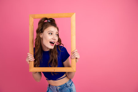 Young woman posing creatively with a wooden frame against a pink background, showcasing trendy fashion and youthful charmの写真素材