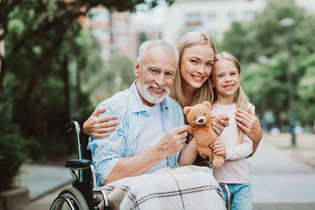 Heartwarming family moment in a sunny park with grandfather in a wheelchair smiling with daughter and granddaughter sharing a teddy bearの写真素材