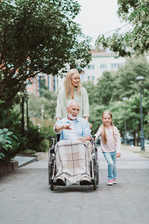 Funny heartwarming family moment grandpa in wheelchair with daughter and granddaughter strolling through park enjoying care connectionの写真素材