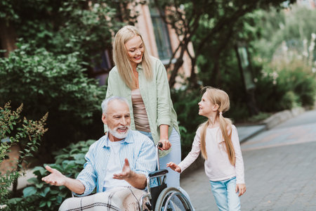 A heartwarming family walk scene with grandpa in a wheelchair being supported by loved ones through a sunny parkの写真素材