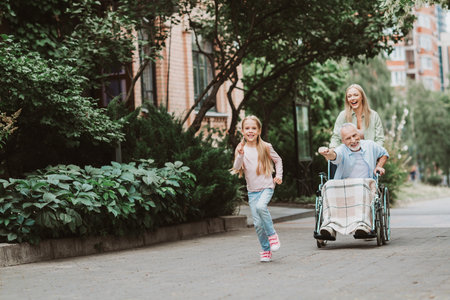 Happy family outing with granddaughter running beside grandfather in a wheelchair pushed by mother outdoors in a green city parkの写真素材