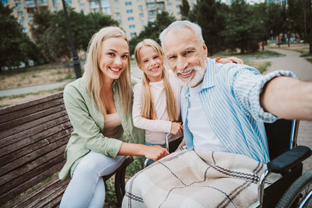 Heartwarming family selfie in park with grandpa in wheelchair smiling with daughter and mom enjoying a sunny dayの写真素材