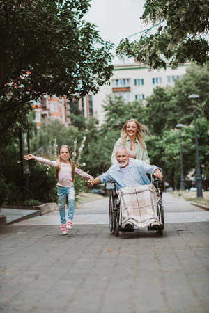 Joyful family outing a grandfather in a wheelchair being pushed by his daughter and a smiling girl...