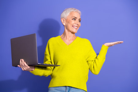 Happy mature woman with short white hair holds a laptop and gestures with her open hand against a purple background.の写真素材