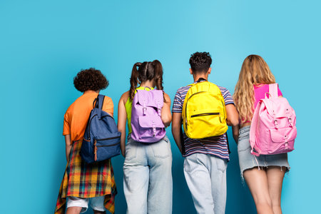Four friends stand with backpacks against a blue wall ready for school and adventure sharing smiles street style fashion and youth energy as they head toward new experiences and memories togetherの写真素材