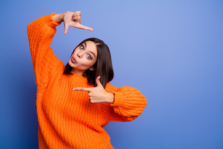 Young woman in vibrant orange sweater striking creative pose with finger frame gesture and playful expression on blue backgroundの写真素材