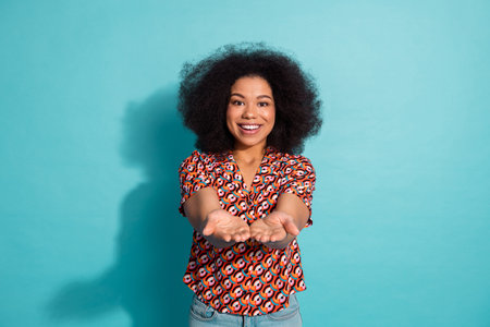 Young woman with voluminous hair in a colorful shirt extends her hands toward the camera against a blue background for lifestyle fashion advertisingの写真素材