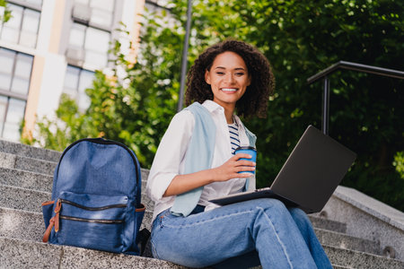 Young woman student outdoor on campus steps with laptop and coffee smiling as she works on a sunny urban day near modern buildingsの写真素材