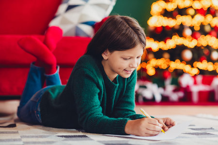 A playful holiday scene of a girl in a green sweater drawing by the Christmas tree filled with warm lights and cozy family vibesの写真素材