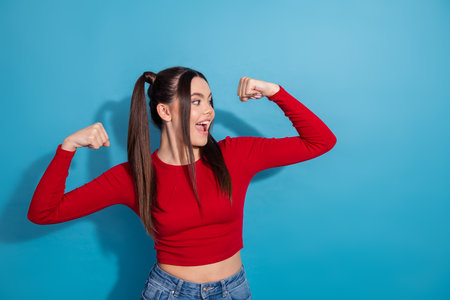 Young woman in red shirt flexing arms and smiling against solid blue background showcasing confidence and styleの写真素材