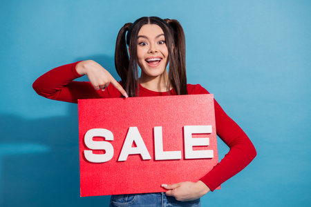 Cheerful young woman holding a sale sign while promoting shopping offers against a vibrant blue backgroundの写真素材
