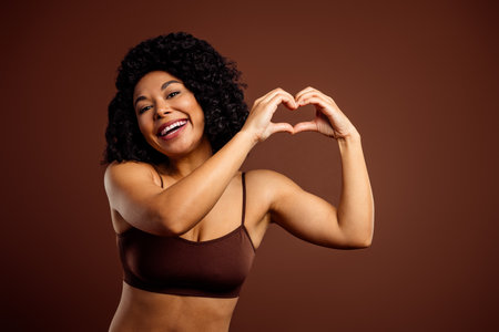Joyful woman with curly hair forming a heart shape with hands on a brown background embodying positivity and confidenceの写真素材