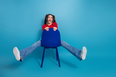 Portrait of a cheerful young woman in a vibrant red top posing playfully with a chair against a bright blue backgroundの写真素材