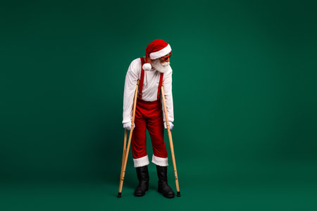 Santa Claus dressed traditionally with crutches against an emerald green background, combining festive cheer with humor.の写真素材