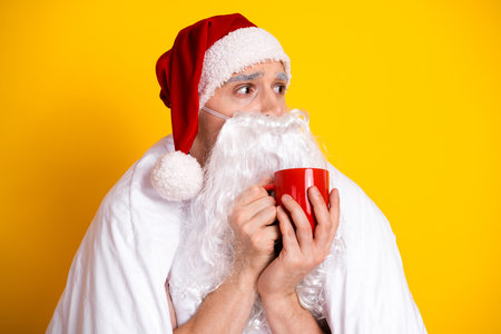 Santa in a red hat with white beard holding a red mug wrapped in a blanket in a bright yellow studio for a cozy Christmas momentの写真素材
