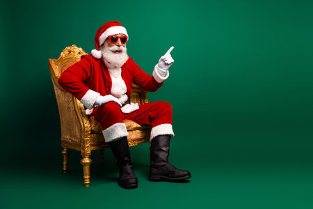 Stylish Santa Claus strikes a festive pose wearing sunglass on a golden chair with a green background, ready for Christmas funの写真素材