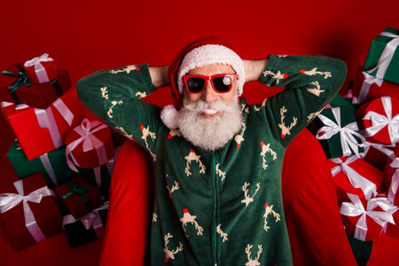 Cheerful Santa in a green Christmas sweater relaxes among gift boxes on a red background delivering holiday joy and festive dreamの写真素材