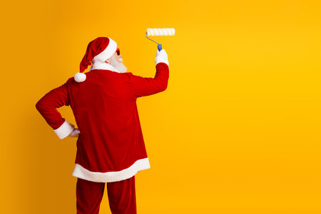 Santa Claus painting a yellow wall with a roller while wearing his traditional red Christmas attire in a vibrant sceneの写真素材