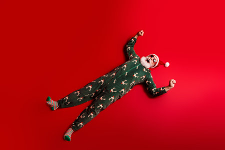 Joyful Santa in Christmas pajamas lies on red background posing for a playful holiday photoの写真素材