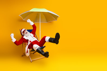 Santa Claus lounging on a deck chair under a sun umbrella against a vibrant yellow background enjoying his timeの写真素材