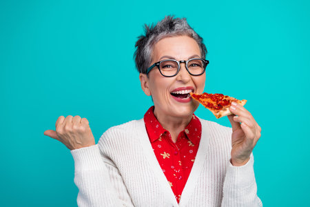 Cheerful elderly woman with glasses enjoys a pizza slice in a teal background wearing a red shirt and white cardigan for a festive holiday stock photoの写真素材