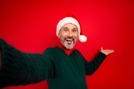Playful Santa selfie with a happy man wearing a green sweater stands against a bold red background spreading Christmas cheer and holiday joy for a festive stock imageの写真素材
