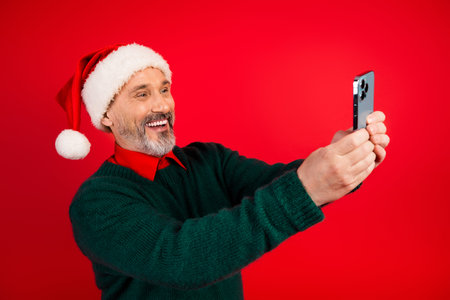 Funny Santa selfie by mature man in green sweater taking joyful christmas photo on red background smiling and posing with smartphone for a holiday season momentの写真素材