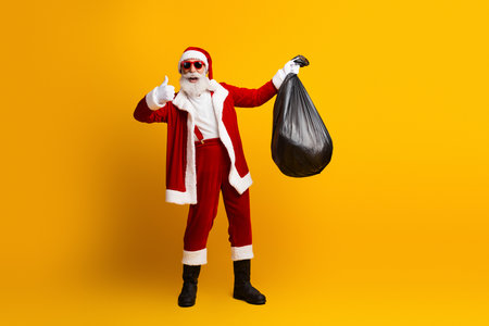 Santa Claus joyfully posing in red outfit holding a bag against a bright yellow backgroundの写真素材