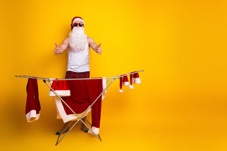 Santa in pajamas gives thumbs up beside a red suit on an ironing board in a bright yellow studio celebrating Christmas humor and homey holiday vibeの写真素材