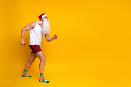 Santa Claus in a white tank top plaid shorts and striped socks runs joyfully in a bright yellow studio settingの写真素材
