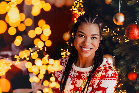 Joyful holiday moment a stylish woman with braids smiles beside a Christmas tree lit with warm lightsの写真素材