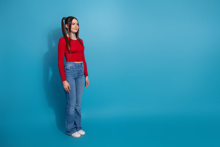 Fashionable young woman posing in casual red top and jeans with vibrant blue background, exuding confidence and stylish eleganceの写真素材
