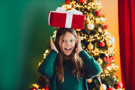 Funny christmas surprise girl balances a red gift on her head beside a twinkling christmas tree at homeの写真素材