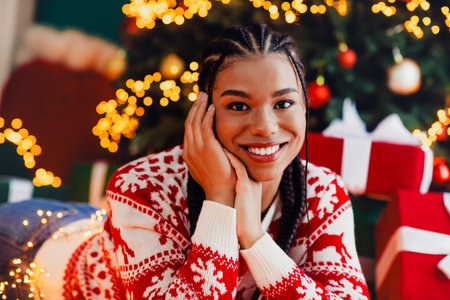 Playful smiling woman in festive christmas sweater relaxing at home with christmas tree lights gifts and warm cozy decorの写真素材