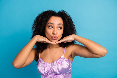 Young woman in lilac satin top poses playfully with hands under chin against bright blue background conveying chic fashion mood and vibrant lifestyleの写真素材