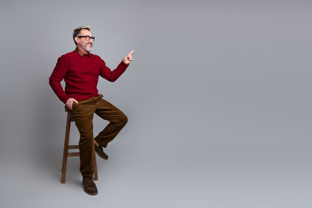 Handsome middle-aged businessman pointing forward seated on stool with gray background wearing glasses and bright casual attireの写真素材