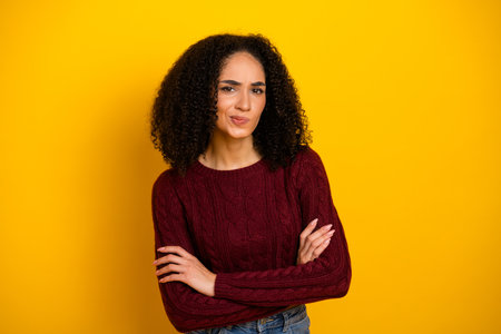 Young mixed race woman with curly hair wearing a burgundy sweater stands against a bright yellow background expressing casual fashion and confidenceの写真素材
