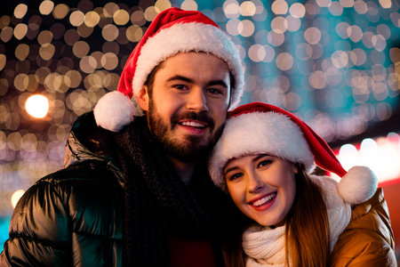 Joyful couple in Santa hats share warm moment under twinkling city lights during festive season smiling at the cameraの写真素材